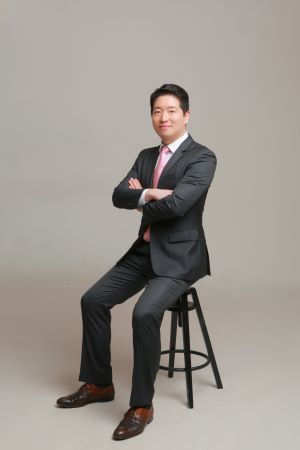 Professional man in a dark suit and pink tie sits on a stool with arms crossed against a neutral backdrop in a business portrait.
