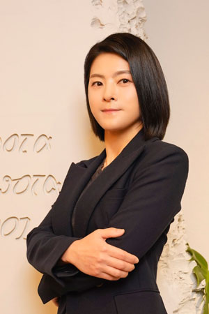 Professional woman with short black hair in a dark blazer, arms crossed, posing against a light textured wall.