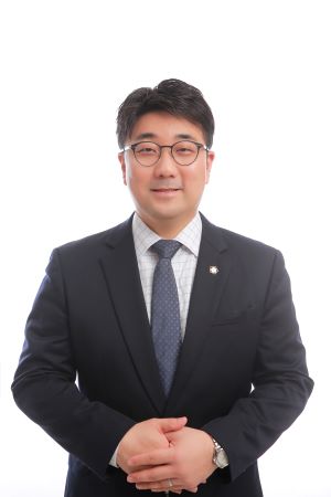 Professional man in a dark suit with a light blue tie and glasses, standing with hands clasped in front and facing the camera.