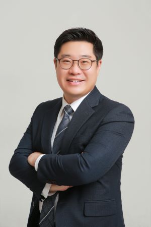 Professional portrait of a smiling Asian man in a navy suit with glasses, arms crossed against a neutral background.