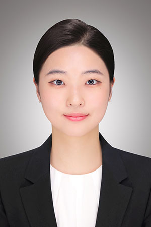 Professional headshot of a young woman in a black blazer and white blouse against a neutral gray background.
