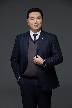 Professional portrait of a man in a navy suit, white shirt, and burgundy sweater, standing with one hand in his pocket against a gray backdrop.