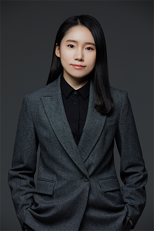 Portrait of a woman with long black hair wearing a dark gray blazer and black shirt, with hands in pockets against a dark backdrop.
