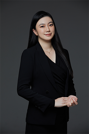 Professional portrait of a woman with long black hair wearing a black blazer and a subtle smile, against a gray backdrop.