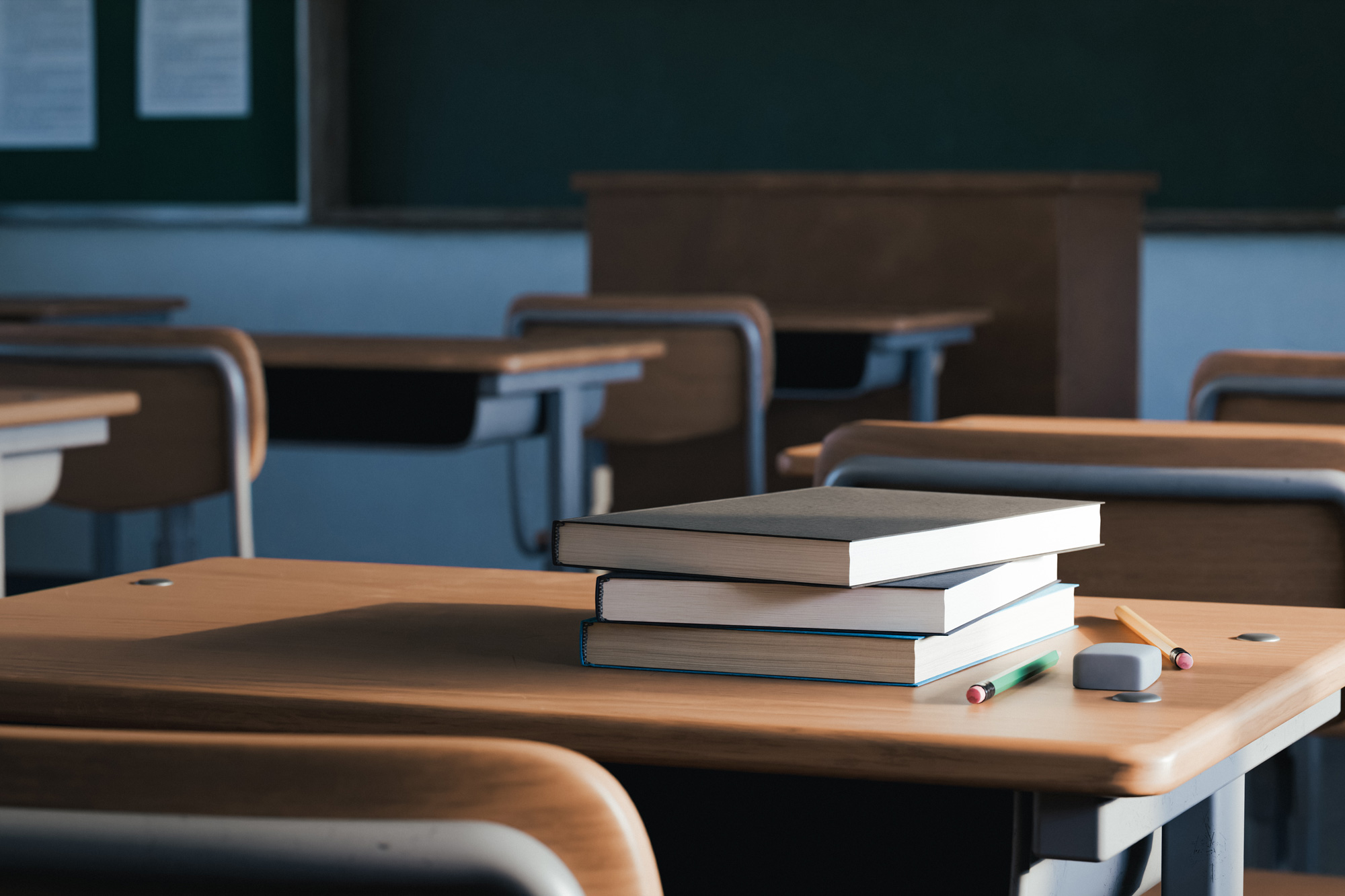 Stack of textbooks on a classroom desk with pencils and an eraser in the foreground; empty desks in the background.