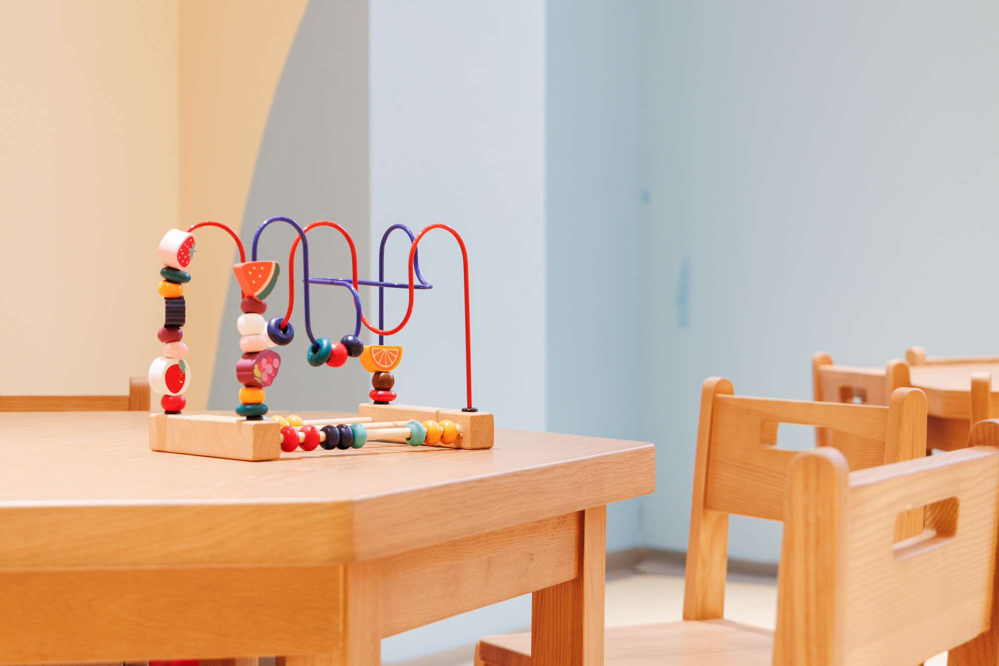 Bead maze toy on a wooden table with colorful beads and red/blue wires in a bright, pastel-colored room with wooden chairs nearby.