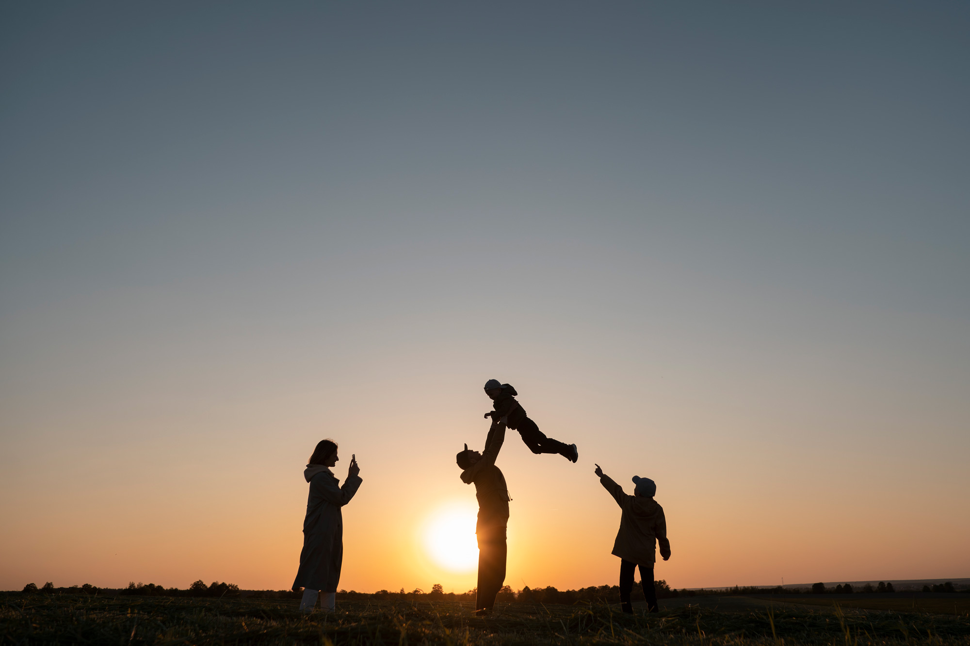 Silhouetted group outdoors at sunset, several people raise their arms while one person lifts a child into the air as the sun sets on the horizon.