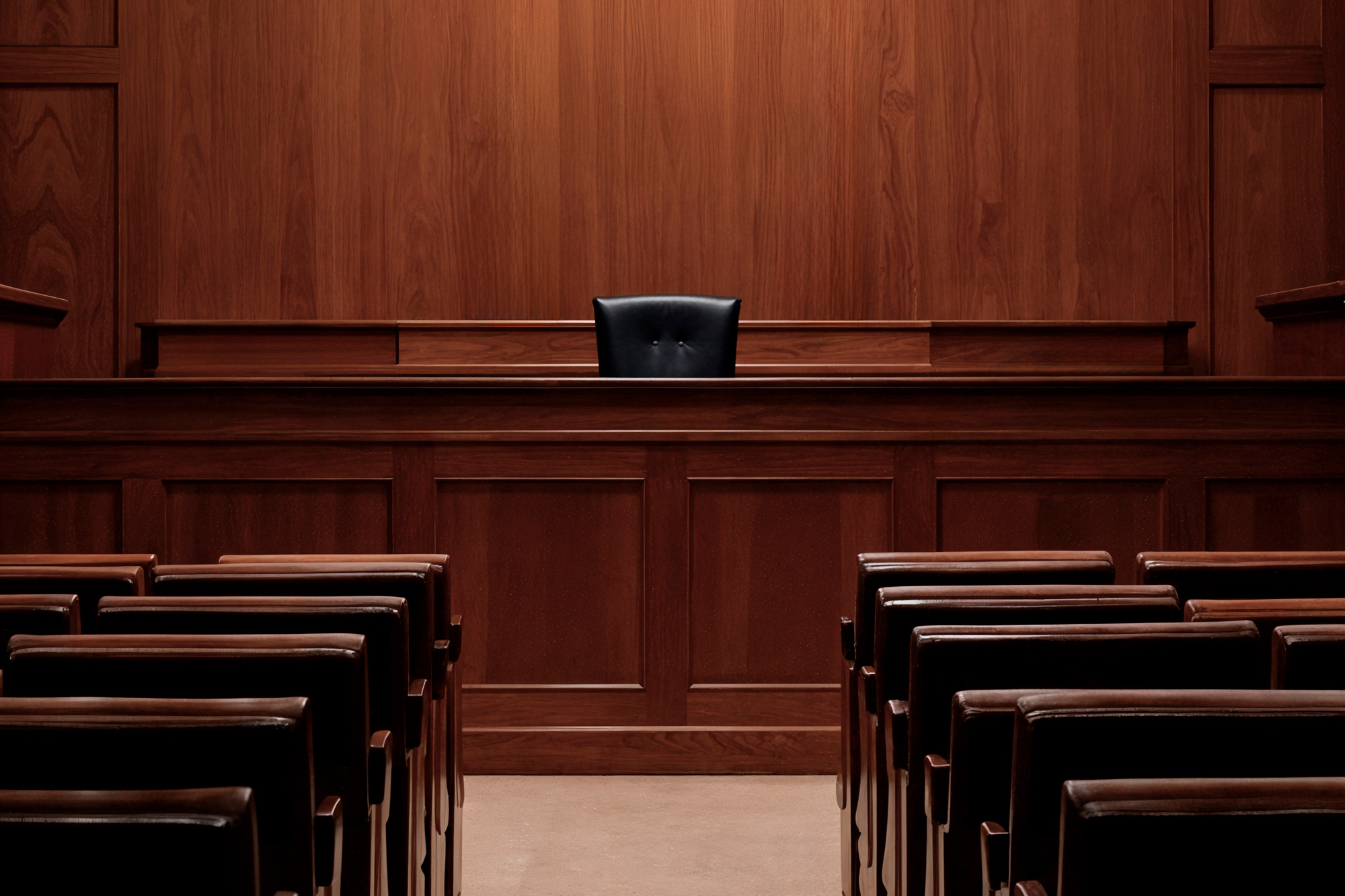 Judge's bench in a wood-paneled courtroom, with a single black leather chair centered behind the bench and empty seating facing forward.
